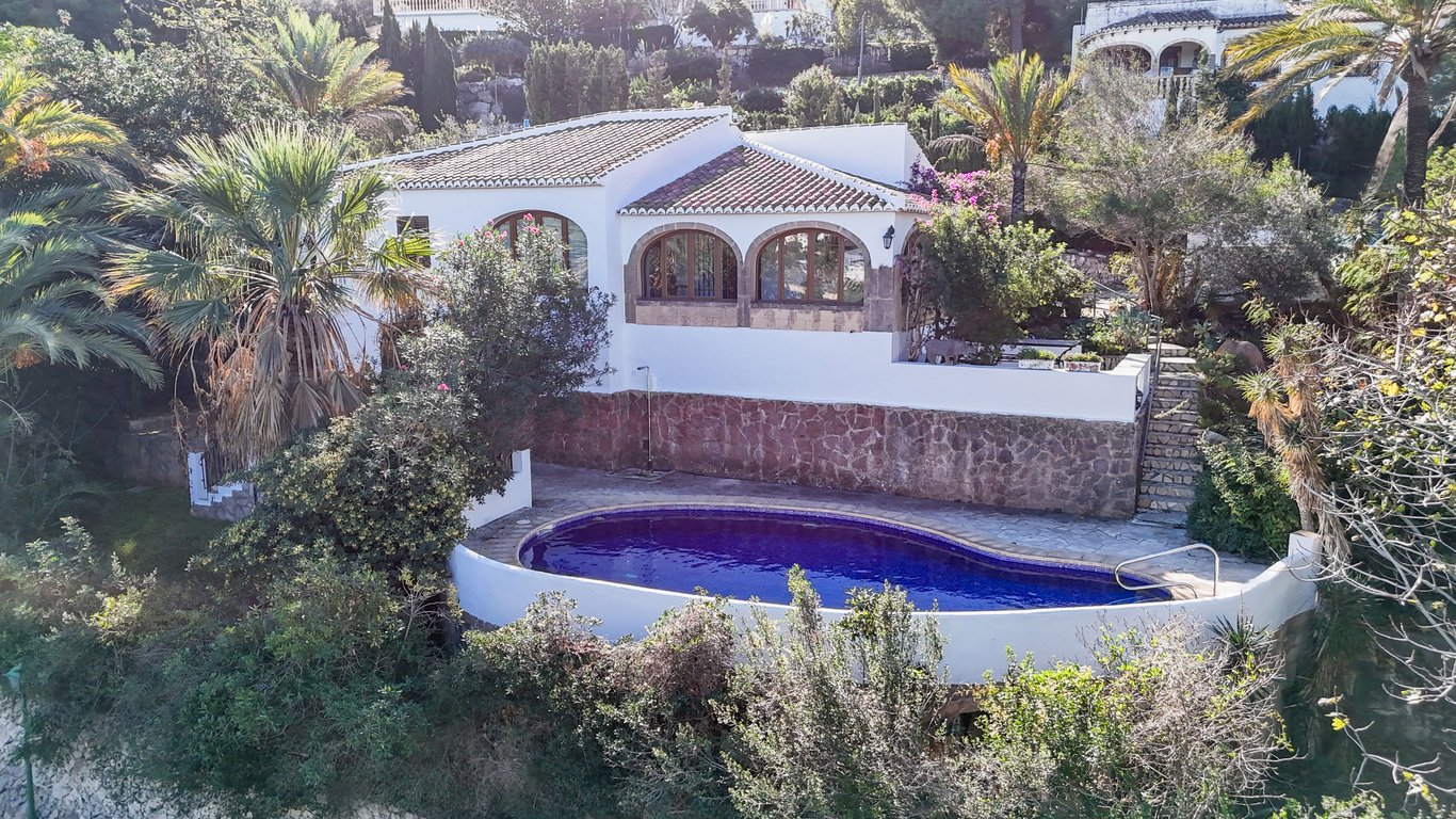 Vista exterior de la villa con piscina privada ovalada, muros de piedra natural y naya con arcos de piedra Tosca rodeada de vegetación mediterránea.