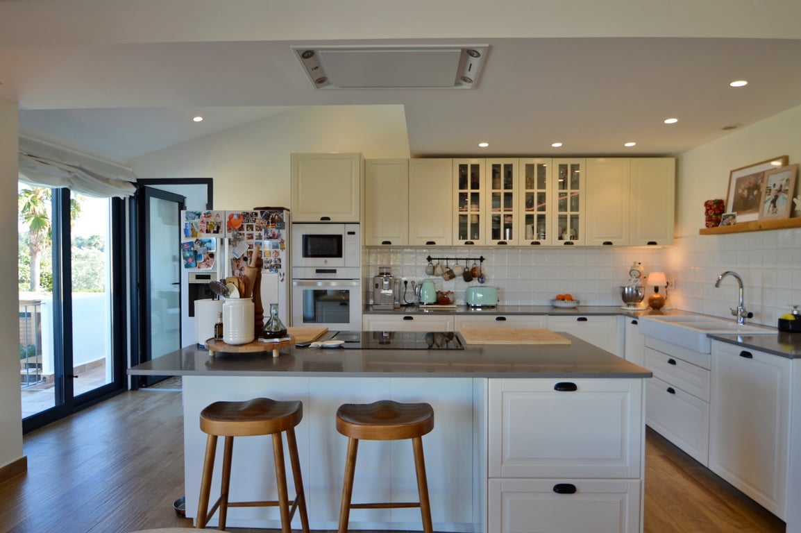 Cocina moderna de concepto abierto con isla central, encimeras grises y taburetes de madera. Incluye electrodomésticos integrados y acceso directo a la terraza exterior.