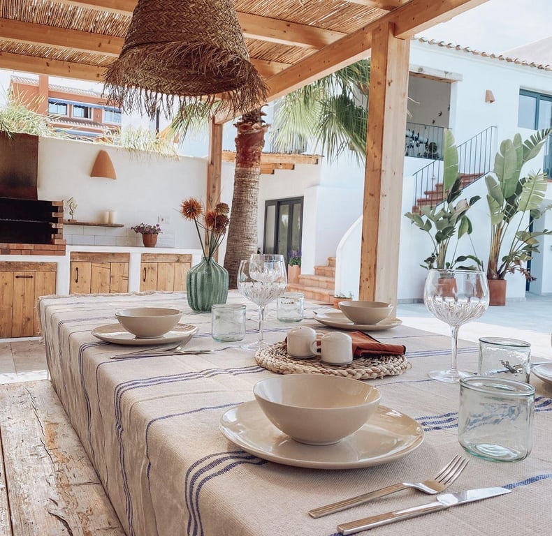 Comedor exterior bajo pérgola de madera y cañizo, con cocina de verano equipada y barbacoa de obra sobre suelos de piedra natural.