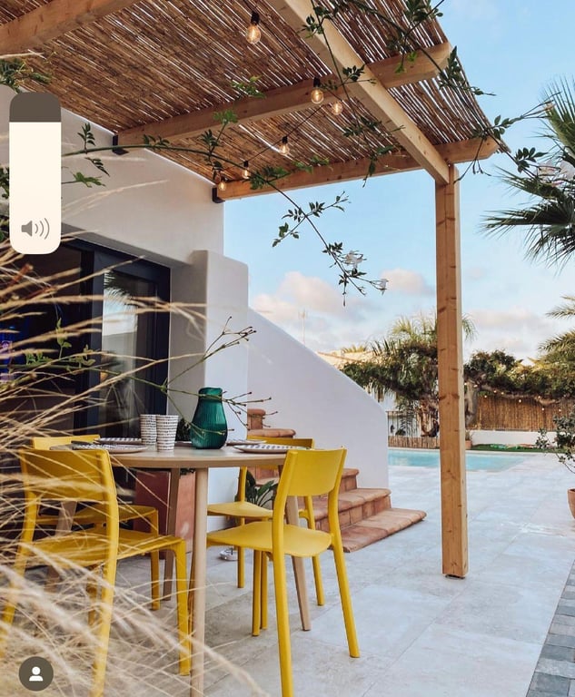 Comedor exterior bajo pérgola de madera y cañizo con guirnaldas de luces. Mobiliario vibrante sobre terraza de piedra clara con vistas a la piscina.