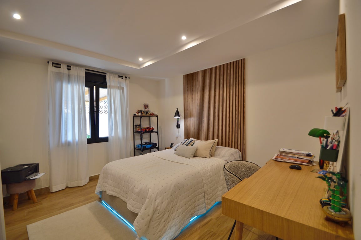 Dormitorio contemporáneo con cabecero de listones de madera, escritorio funcional y cama con iluminación LED inferior. Presenta suelos cálidos y techo con luces encastradas.