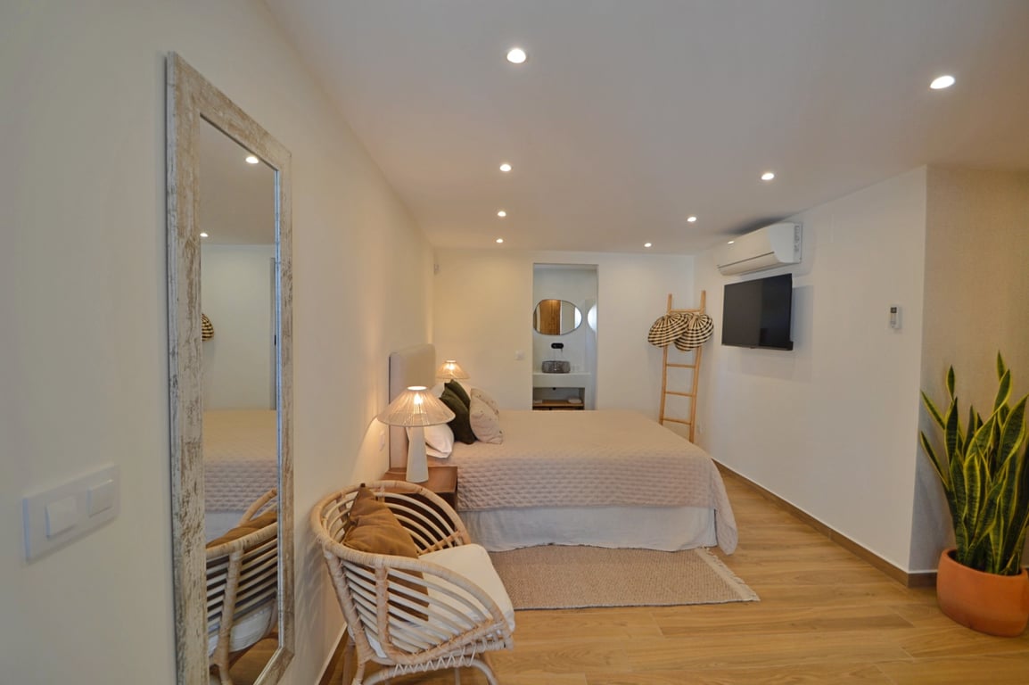 Dormitorio de estilo ibicenco con suelos de madera, iluminación LED empotrada, aire acondicionado y acceso directo a un baño integrado.