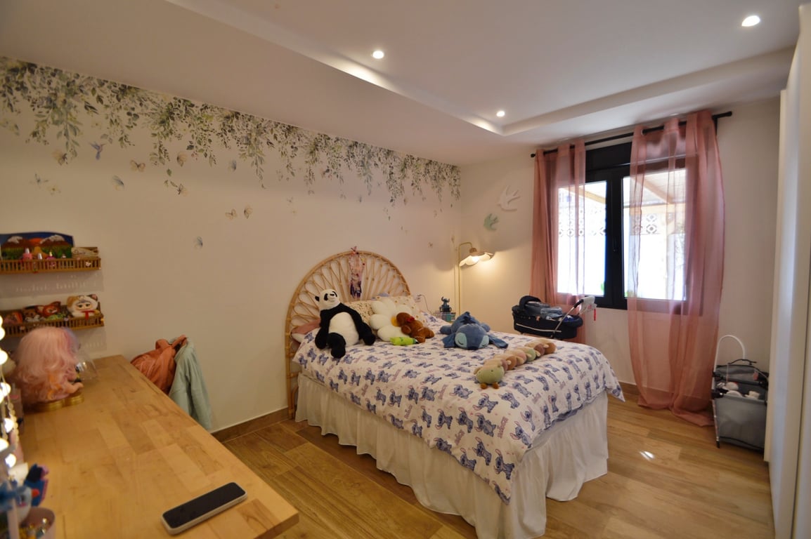 Dormitorio luminoso con cabecero de mimbre, mural botánico y suelos de madera. Incluye iluminación empotrada y luz natural mediante ventanal con cortinas sutiles.