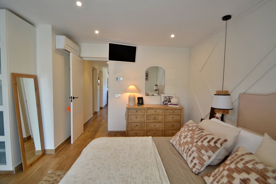 Dormitorio luminoso con suelos de madera, pared decorativa con molduras geométricas, aire acondicionado individual y mobiliario de madera natural que aporta calidez.