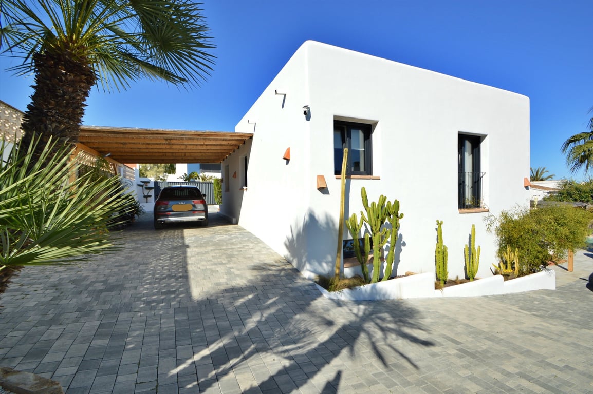 Fachada blanca de estilo ibicenco con carport de vigas de madera, amplia zona de aparcamiento pavimentada y jardín mediterráneo con cactus.