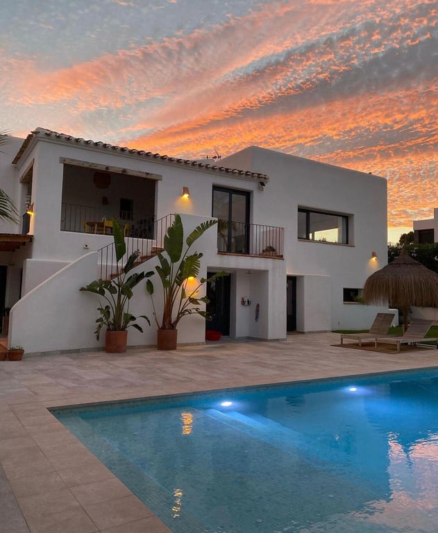 Fachada de estilo ibicenco al atardecer con piscina de agua salada iluminada, amplia terraza de piedra y vegetación mediterránea bajo un cielo vibrante.