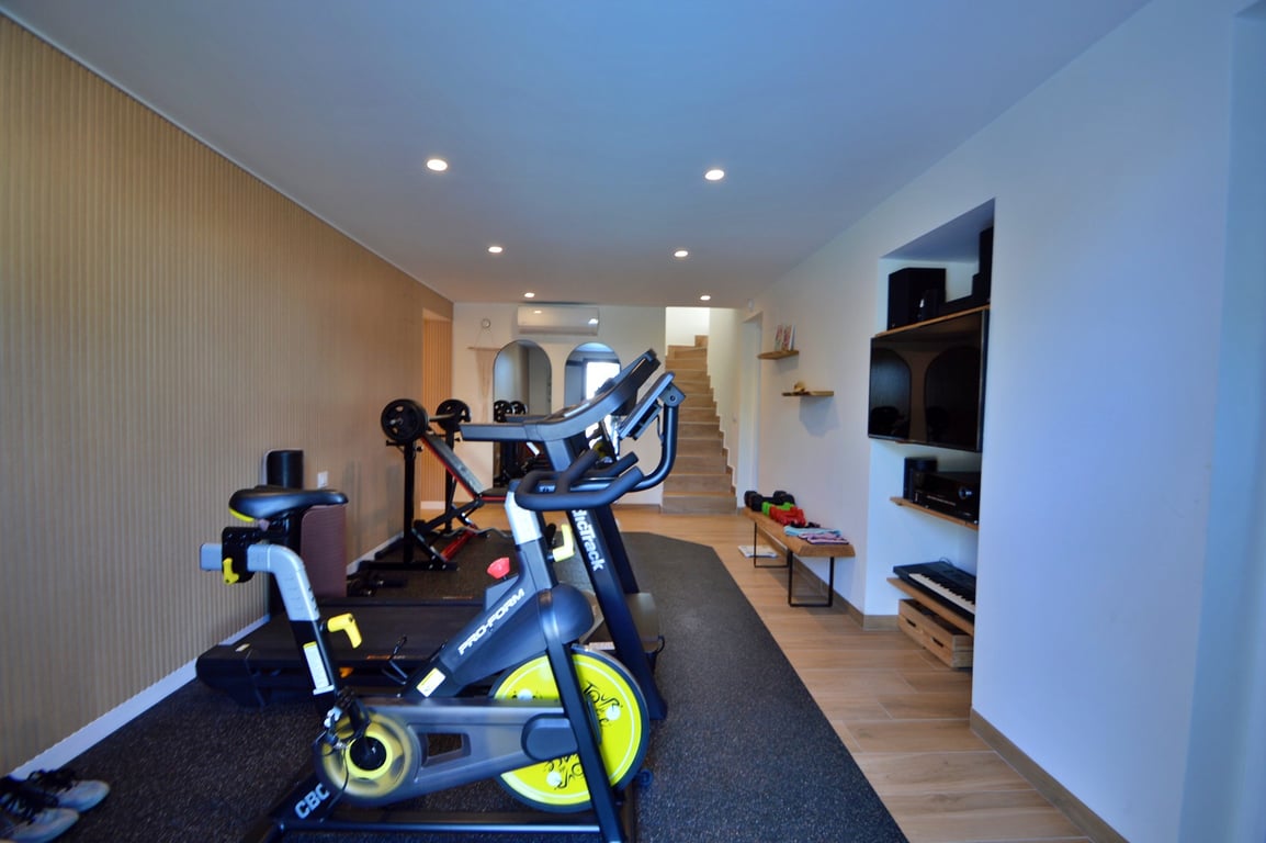 Gimnasio privado equipado con bicicleta de spinning, cinta de correr y pesas, con pared panelada en madera, iluminación empotrada y zona multimedia integrada.