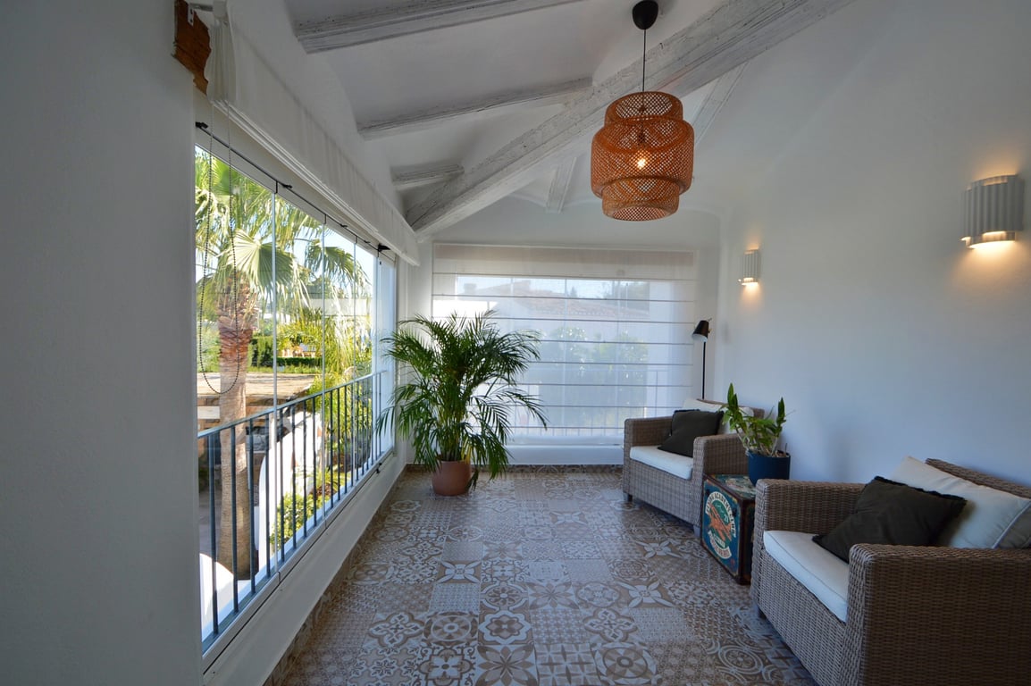 Naya acristalada con vigas de madera blancas, suelo de baldosa hidráulica decorativa y mobiliario de mimbre con vistas al jardín mediterráneo.