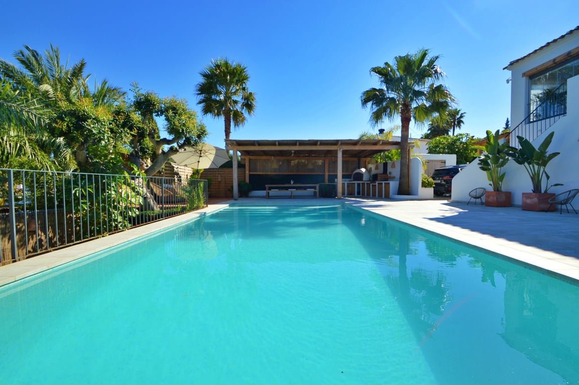 Piscina rectangular rodeada de una amplia terraza soleada, con zona de comedor exterior bajo pérgola de madera y jardín mediterráneo con palmeras.