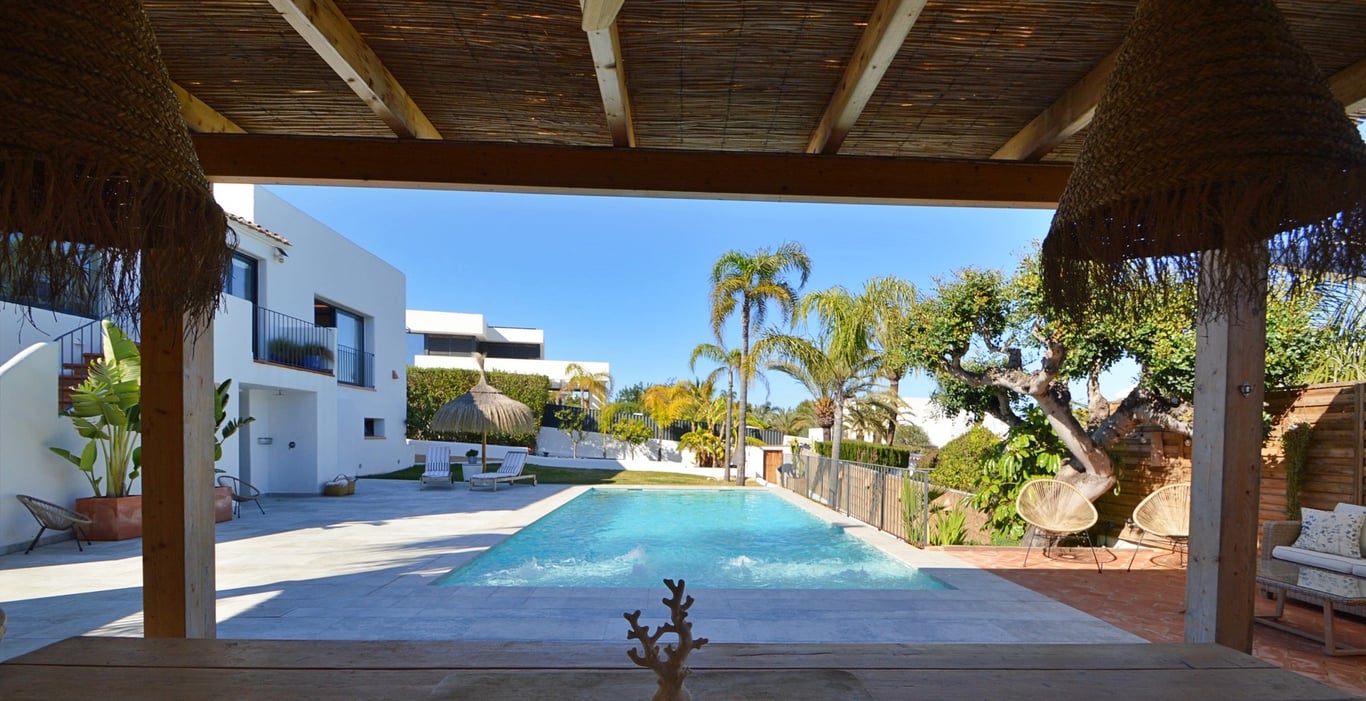 Vista desde el porche sombreado con techo de cañizo hacia la piscina de agua salada y el jardín mediterráneo con palmeras.