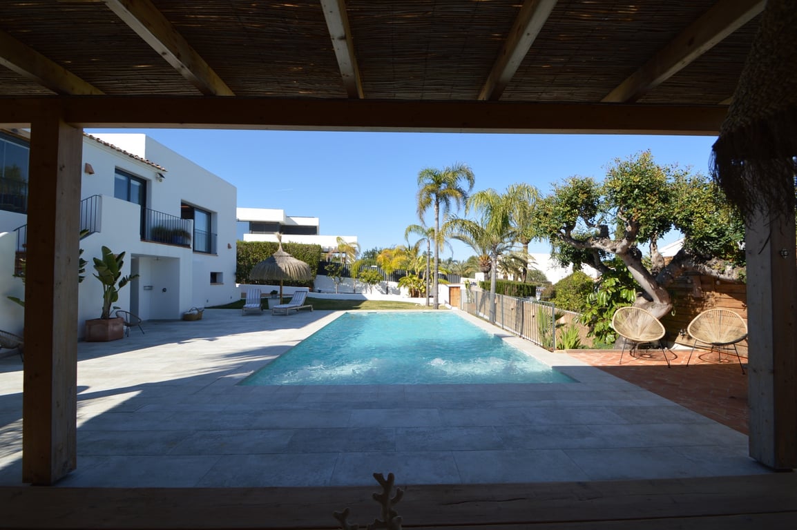Vista desde la pérgola de madera hacia la piscina de agua salada, rodeada de amplias terrazas de piedra clara y jardines mediterráneos con palmeras.