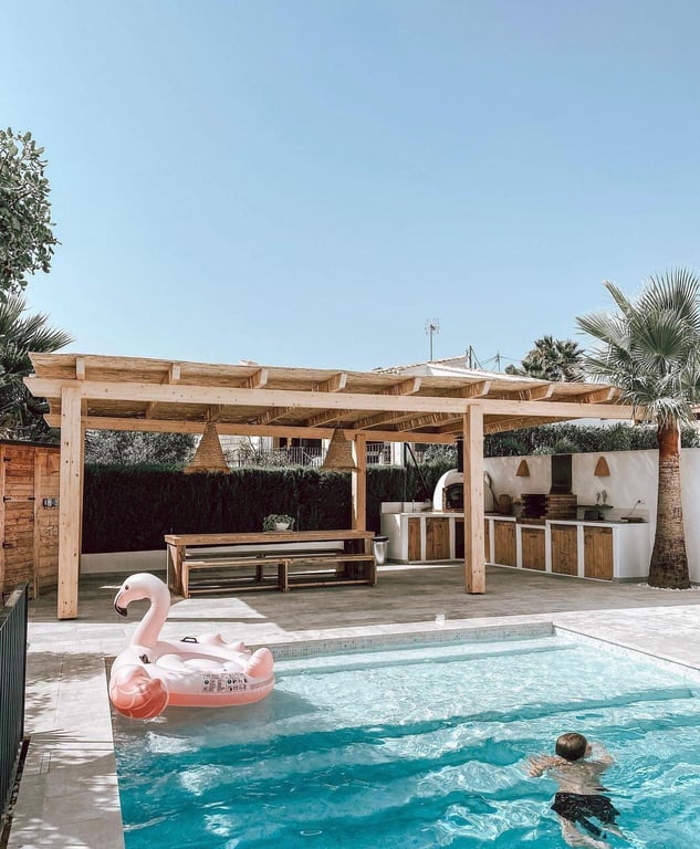 Zona de comedor exterior y cocina de verano con horno de pizza y barbacoa, protegida por una pérgola de madera frente a la piscina de agua salada.