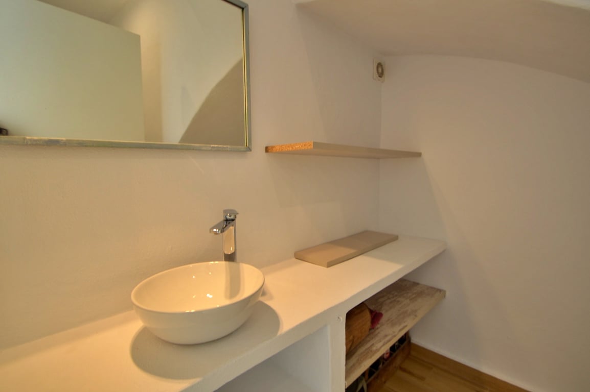 Baño de diseño minimalista con lavabo sobre encimera blanca, estanterías integradas y espejo rectangular. Paredes blancas y detalles en madera aportan calidez al espacio.