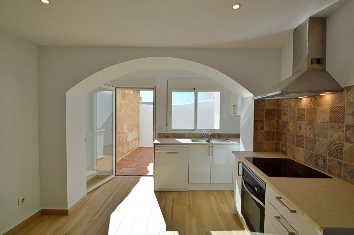 Cocina moderna integrada con arco arquitectónico, encimera clara, azulejos decorativos y acceso directo a un patio privado con suelos de terracota.