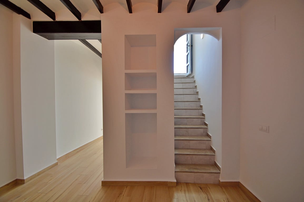 Distribuidor interior con vigas de madera vista, estantería integrada en la pared y escalera de gres que conduce a la planta superior bajo luz natural.
