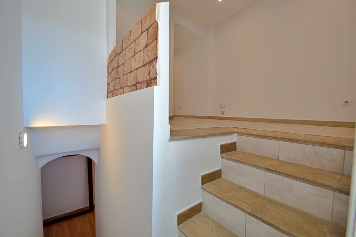 Escalera interior con peldaños cerámicos de efecto madera, paredes blancas y un detalle decorativo de ladrillo visto que separa los niveles.