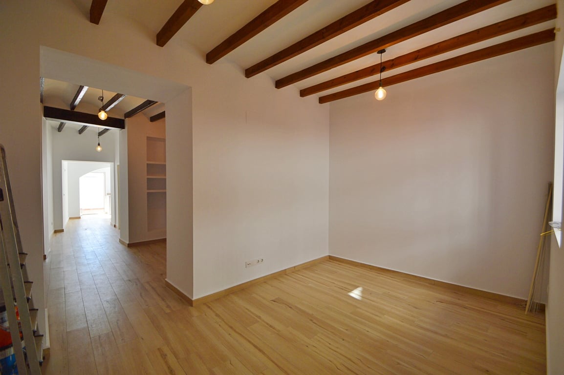 Estancia diáfana con vigas de madera vista, suelos de tarima clara y paredes blancas. La iluminación colgante resalta el diseño tradicional renovado y la amplitud del espacio.