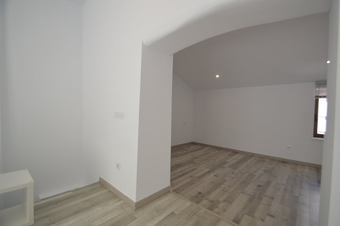 Habitación renovada con suelos de tarima clara, paredes blancas y focos empotrados. El espacio incluye un acceso abierto y una ventana con marco de madera.