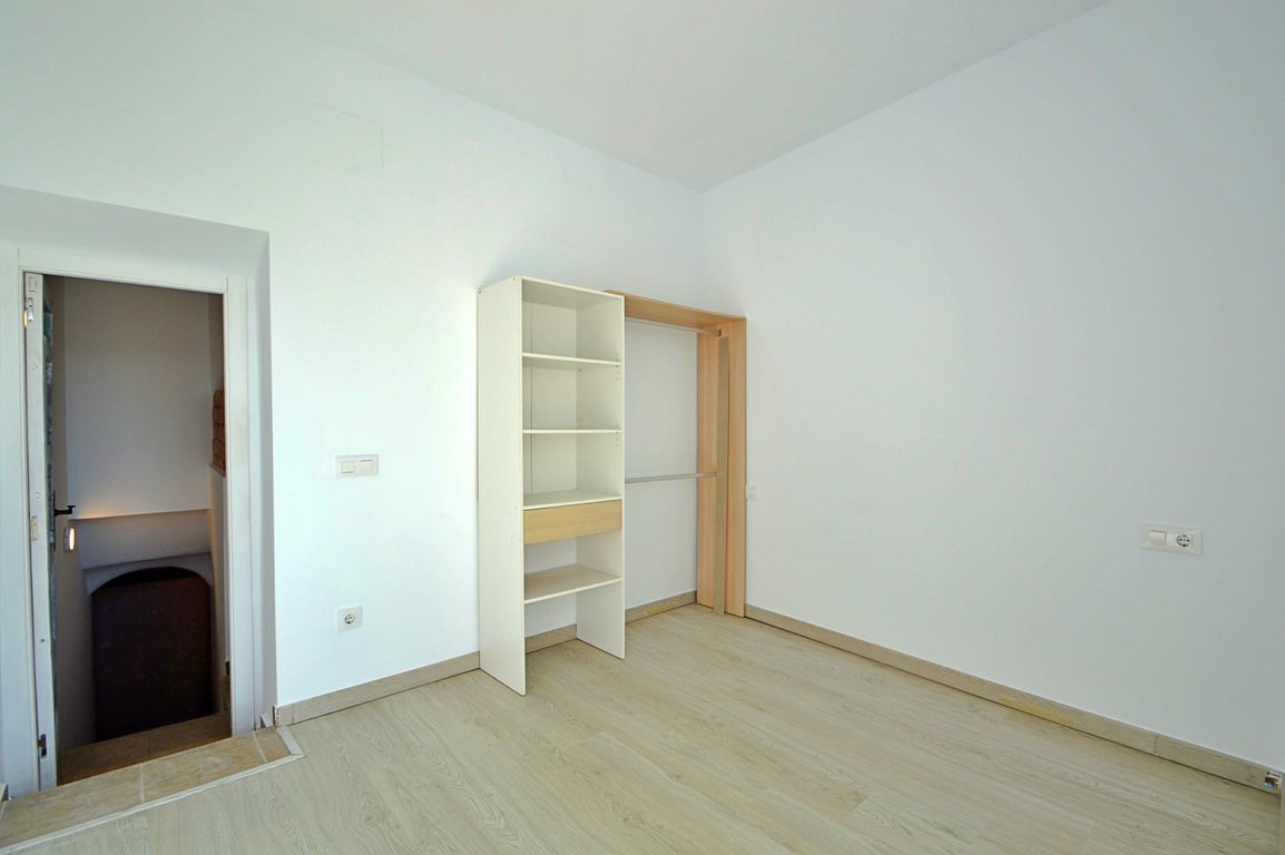 Habitación versátil con suelos de gres efecto madera, paredes blancas y estantería abierta con zona de colgado, situada junto al acceso de la escalera.