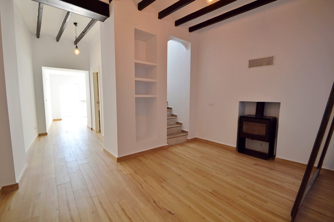 Salón con vigas de madera oscura, suelos laminados claros y chimenea de hierro integrada. Incluye estanterías de obra y acceso a la escalera de la planta superior.