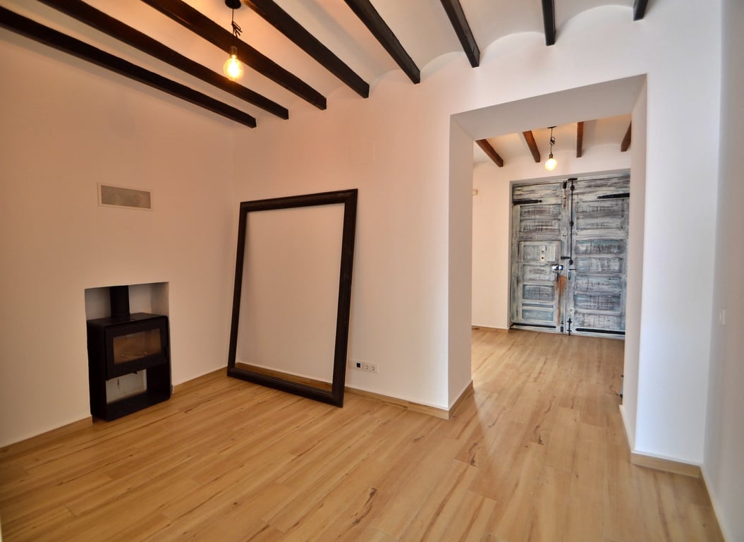 Salón con vigas de madera vistas, estufa de leña encastrada y suelos claros. Al fondo destaca un portón rústico restaurado que aporta carácter histórico.