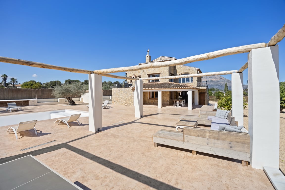 Amplia terraza exterior con pérgola de vigas de madera, zona de estar y vistas a la fachada de piedra de la villa con el Montgó al fondo.