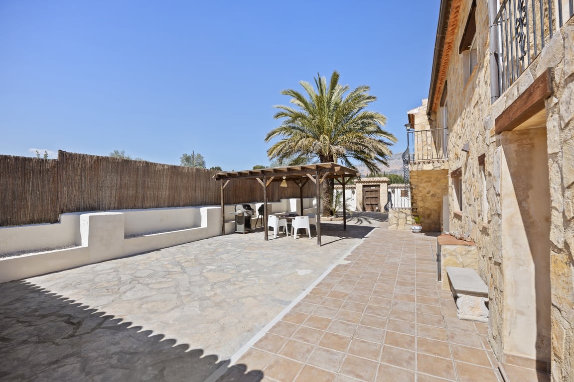 Amplia terraza exterior con suelo de piedra y baldosas, zona de pérgola de madera con barbacoa y fachada de piedra natural bajo un cielo despejado.