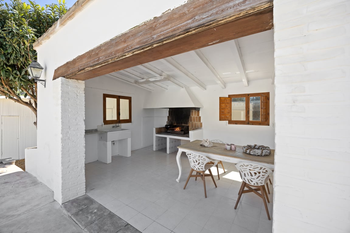 Cocina exterior cubierta con barbacoa de obra, fregadero de piedra y zona de comedor bajo un techo de vigas blancas y un robusto dintel de madera.