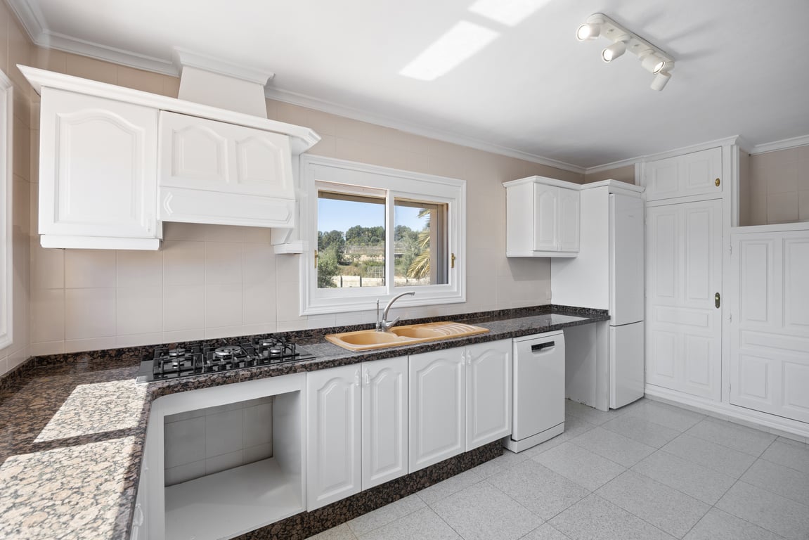 Cocina funcional con armarios blancos, encimera de granito oscuro y fregadero de doble seno. Incluye placa de gas, campana clásica y ventana con vistas al entorno mediterráneo.