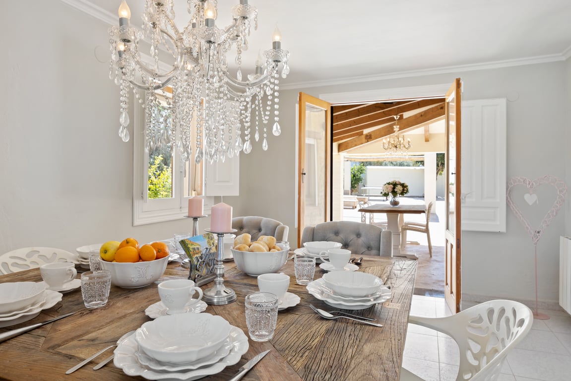 Comedor luminoso con mesa de madera rústica y lámpara de araña de cristal, con acceso directo a la naya cubierta mediante puertas de madera.