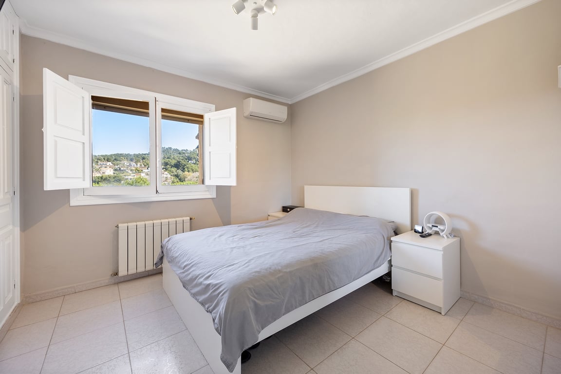 Dormitorio luminoso con contraventanas blancas, aire acondicionado y vistas despejadas al entorno de Jávea. Incluye suelos cerámicos y tonos neutros.