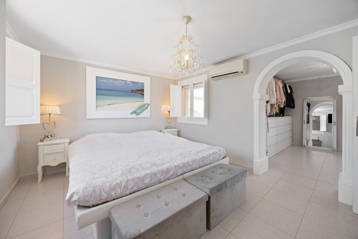 Dormitorio principal con vestidor integrado tras un arco decorativo, suelos cerámicos, aire acondicionado y ventanas con contraventanas de madera blanca.