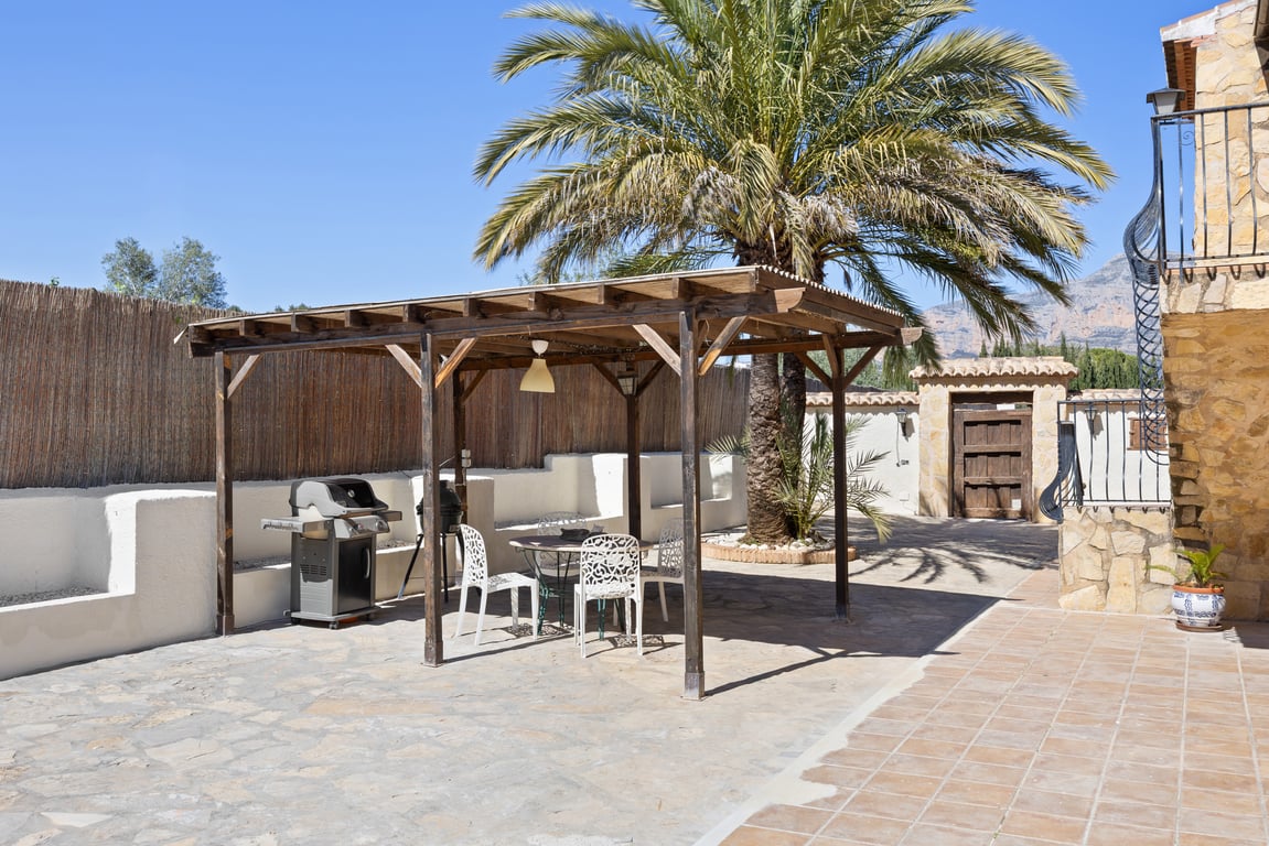 Pérgola de madera con zona de comedor exterior y barbacoa, situada en una terraza de piedra junto a una gran palmera y muros de piedra natural.