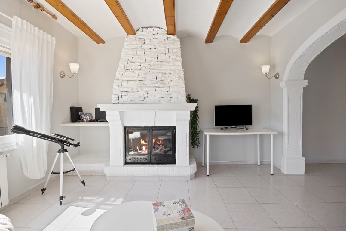 Salón con chimenea de piedra blanca, vigas de madera vistas en el techo y un arco de piedra tradicional que conecta los espacios.
