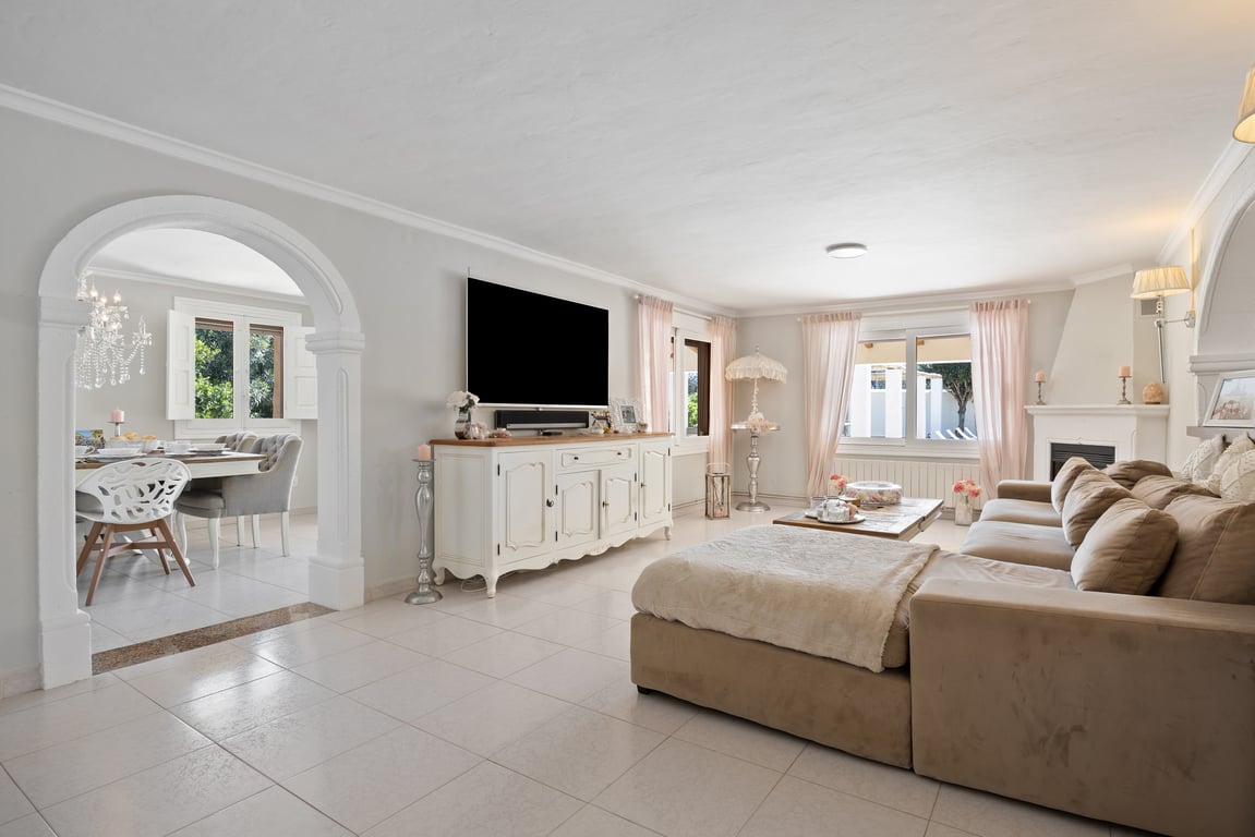 Salón luminoso con suelos cerámicos, sofá rinconero y chimenea. Un arco decorativo conecta con el comedor, destacando la amplitud y la luz natural de la estancia.
