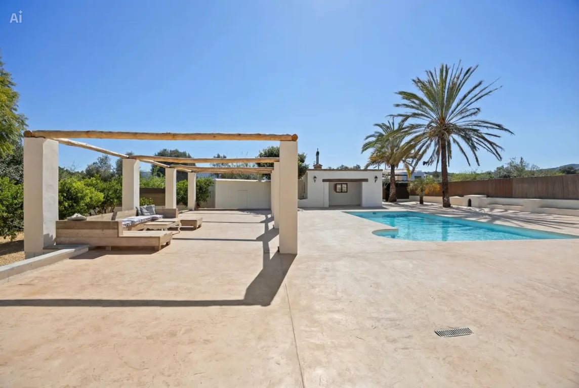 Zona de piscina con amplia terraza pavimentada, pérgola de vigas de madera rústica y zona de estar integrada junto a palmeras y jardines mediterráneos.