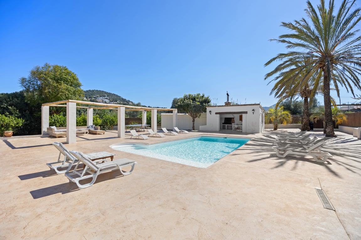 Zona de piscina de 5x10m con amplia terraza pavimentada, cocina exterior equipada, pérgola con vigas de madera y entorno mediterráneo con palmeras.