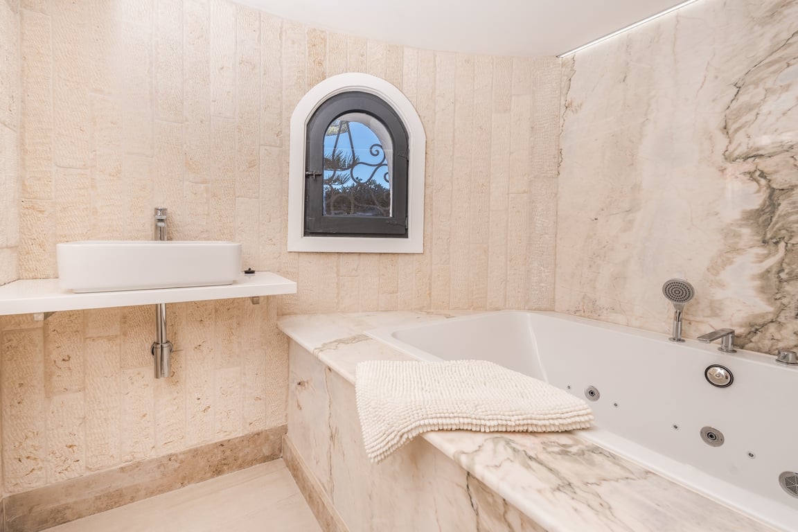 Baño con bañera de hidromasaje integrada en mármol, lavabo sobre encimera y paredes revestidas en piedra natural con ventana de arco característica.