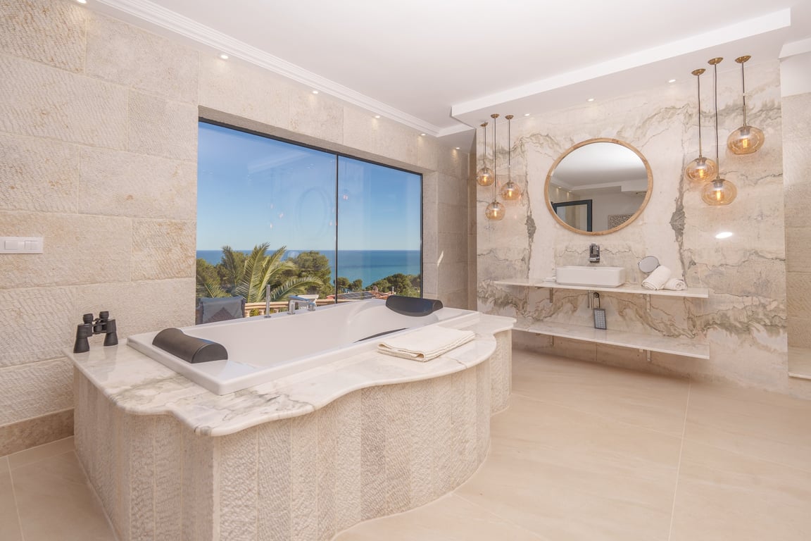 Baño de la suite principal con bañera encastrada en mármol, paredes de piedra caliza y ventanal con vistas despejadas al mar Mediterráneo.