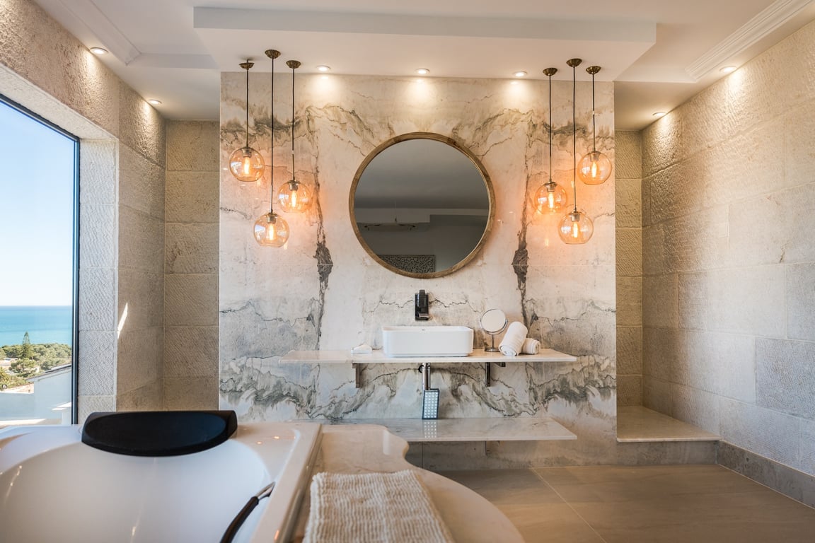 Baño de la suite principal con pared de mármol veteado, lavabo sobre encimera flotante, luminarias colgantes de diseño y vistas despejadas al mar Mediterráneo.