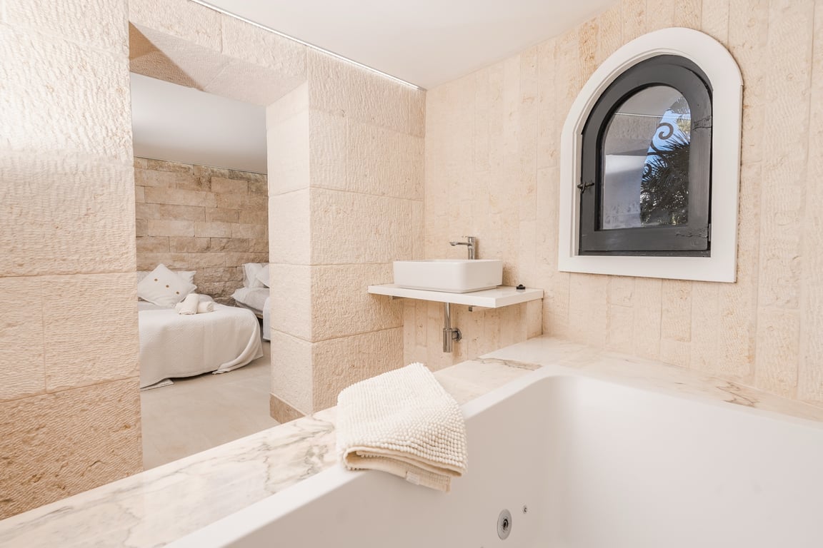 Baño integrado con paredes de piedra natural, bañera con borde de mármol y lavabo de diseño, conectado visualmente con el dormitorio a través de un arco de piedra.