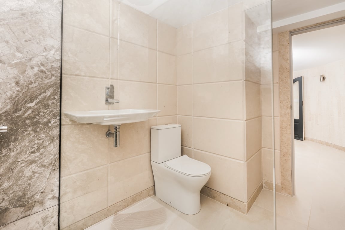 Baño moderno con revestimiento de piedra caliza en tonos crema, inodoro de diseño actual y lavabo suspendido. Incluye mampara de cristal y acabados en mármol veteado.