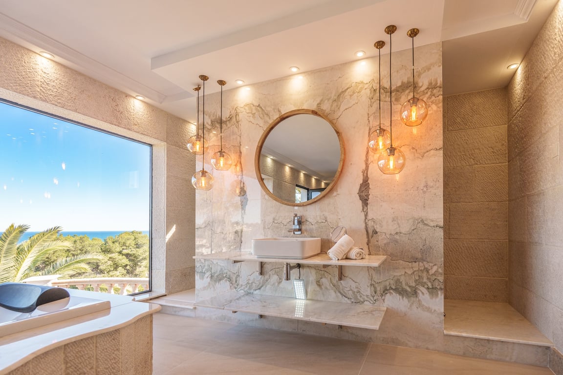 Baño principal con paredes de mármol veteado, lavabo sobre encimera flotante y espejo circular. Destacan las lámparas colgantes de cristal y las vistas al mar desde el gran ventanal.