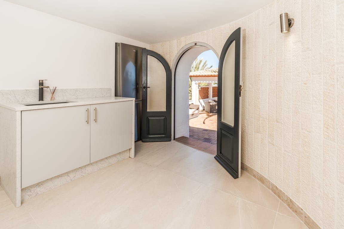 Cocina compacta con encimera de mármol y paredes de piedra natural, conectada a la terraza exterior mediante un arco con puertas dobles oscuras.