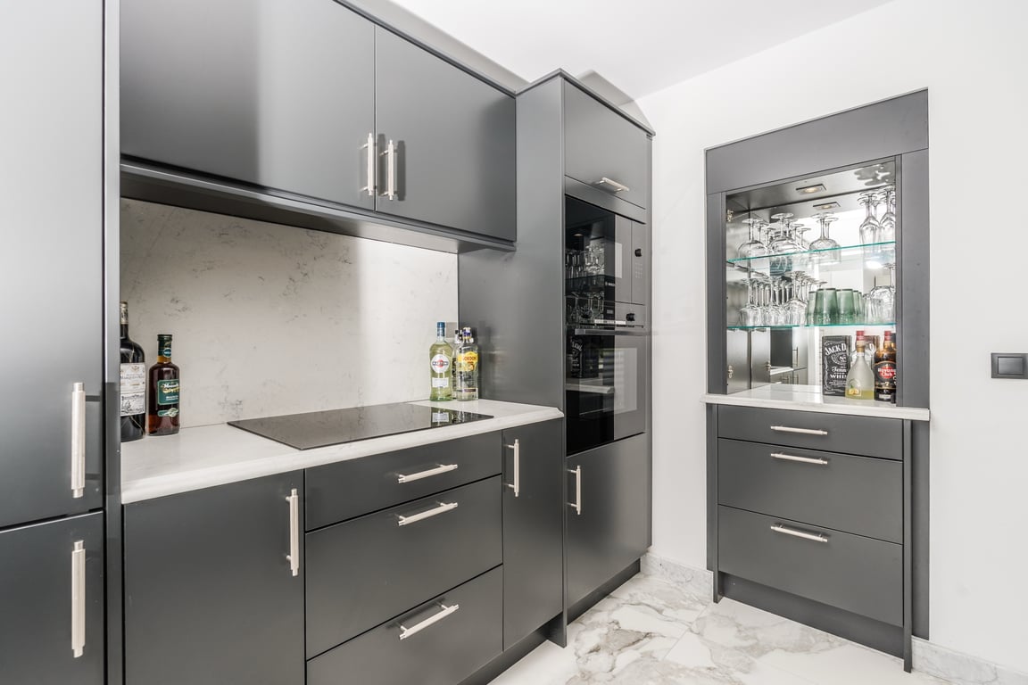 Cocina moderna con armarios gris antracita, encimera de mármol blanco y electrodomésticos integrados. Incluye un mueble bar con vitrina iluminada y suelos de mármol veteado.