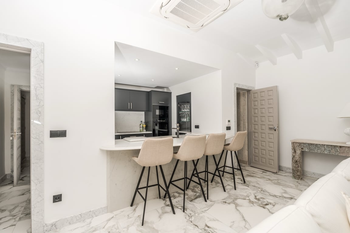 Cocina semiabierta con barra de mármol y taburetes modernos. Destacan los suelos de mármol veteado, marcos de piedra y techos con vigas blancas.