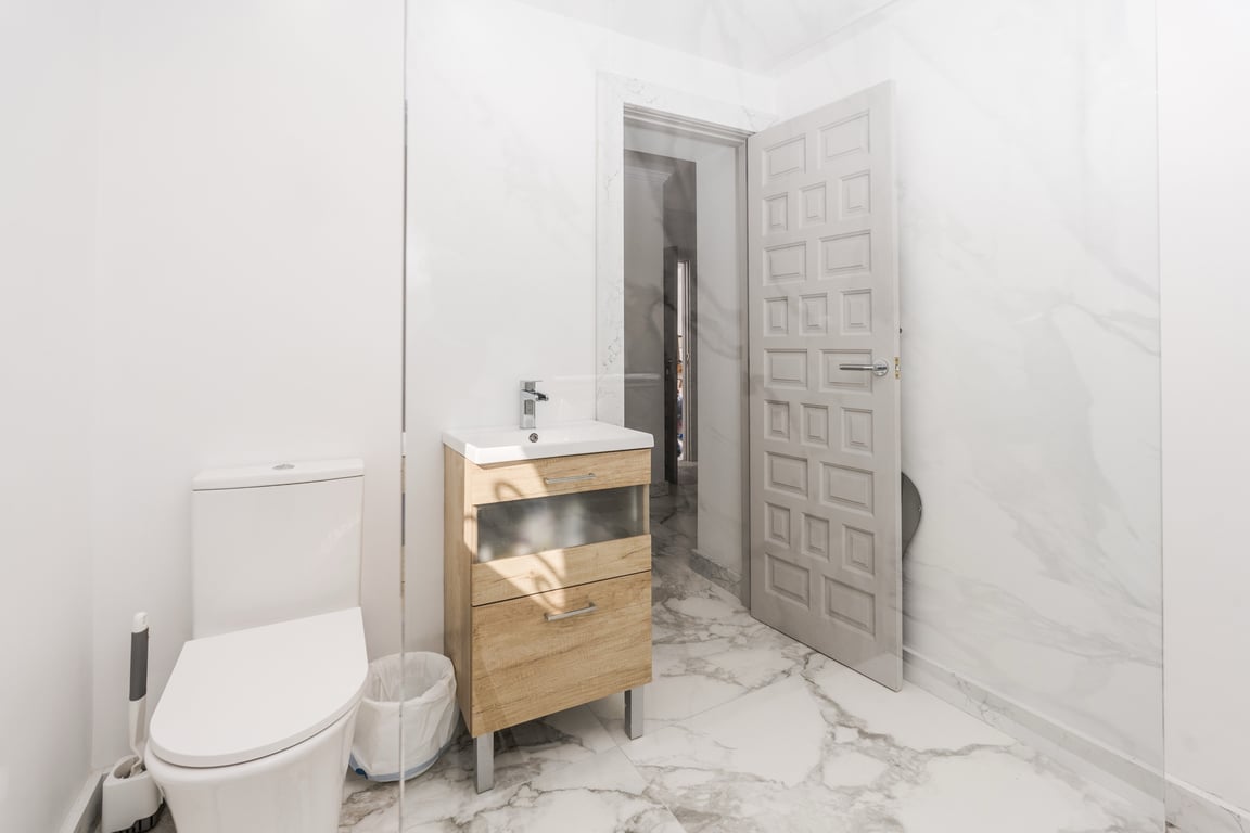 Cuarto de baño moderno con revestimiento de mármol blanco, mueble de lavabo en madera clara y mampara de cristal fija que aporta luminosidad.