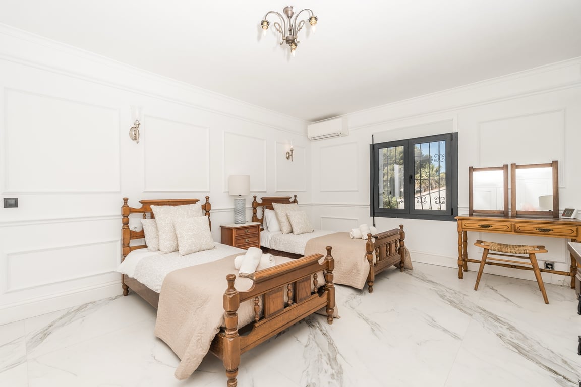 Dormitorio con dos camas individuales de madera tallada, suelos de mármol veteado y paredes con molduras clásicas. Incluye un tocador de madera y aire acondicionado.