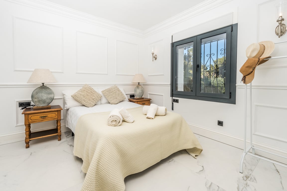 Dormitorio luminoso con paredes decoradas con molduras, suelos de mármol blanco y ventana de carpintería oscura. Incluye mesitas de noche de madera y lámparas con base de piedra.