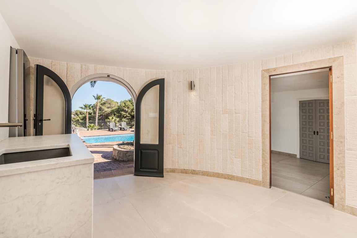 Espacio interior con paredes de piedra natural texturizada y acceso directo a la piscina mediante un arco. Incluye encimera de mármol con fregadero integrado y suelos pulidos.